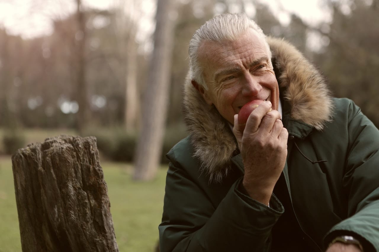 Man in Green Parka Jacket Covering His Face With His Hand Eating an Apple; represents the concept of healthy nutrition for diabetes diet for older adults