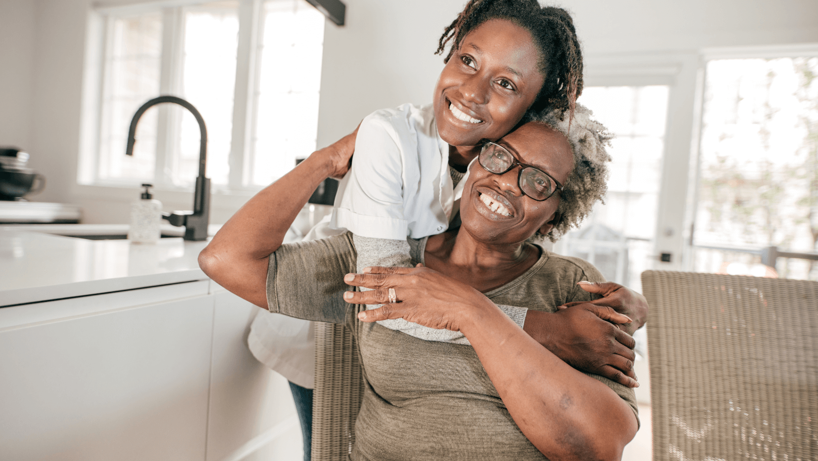 A younger Black woman hugs an older Black woman. They smile and look ahead. They are in a clean room that is modified to be safer for seniors.